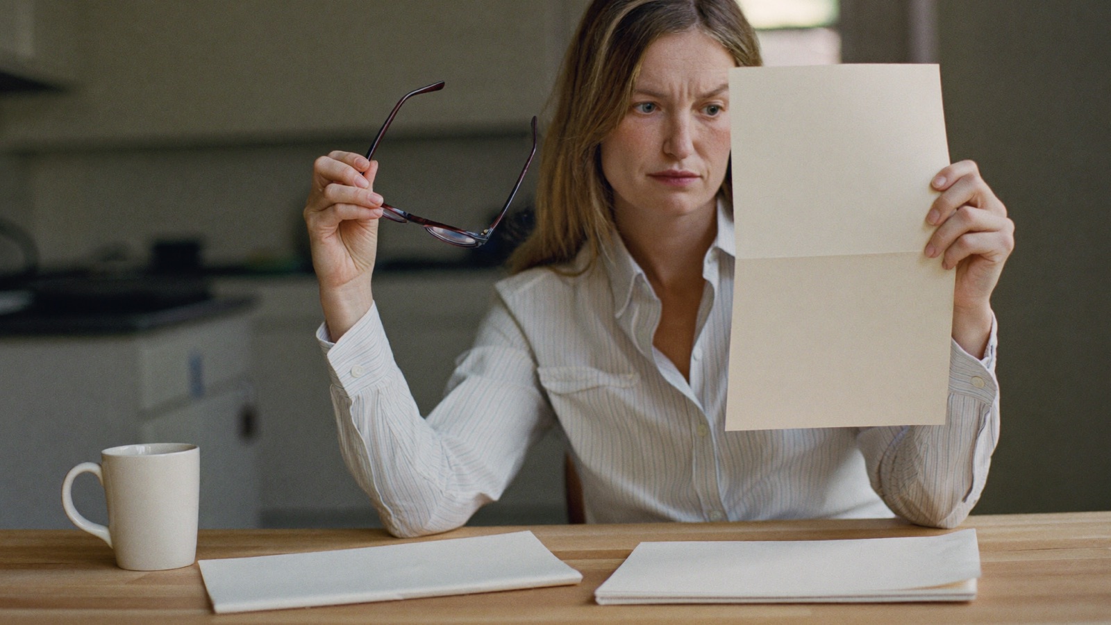 A small-business owner at a kitchen table holding up one agency proposal with two more folded on the table in front of her, glasses held in one hand, looking unconvinced