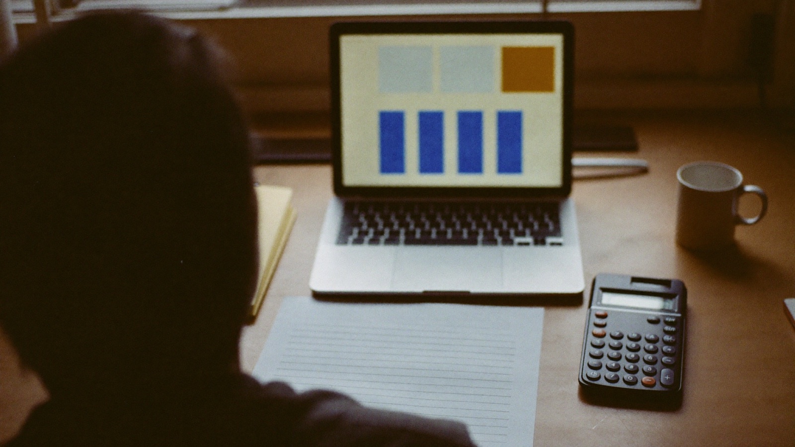 A small-business owner at a desk comparing an advertising dashboard on a laptop with a printed profit-and-loss sheet and a calculator