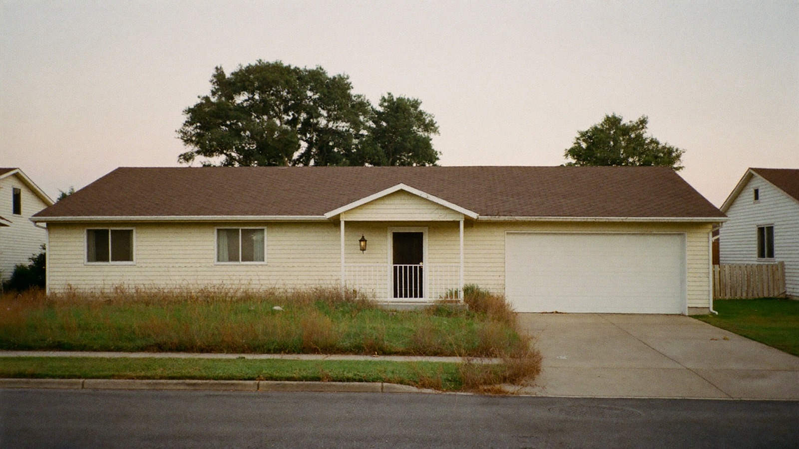 A modest single-story suburban house with faded siding and an overgrown lawn, photographed from across the street in late afternoon light